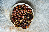 Top view of fresh chestnuts placed in ceramic bowl with dried orange slices in a concrete background