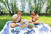 A happy couple sits on a picnic blanket enjoying food and each other's company in a sunlit park with trees in the background.