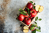 Top view of whole fresh red ripe organic delicious apples with green leaves filled in wooden box with half cut pieces and placed on gray surface in daylight