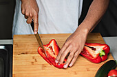 Concentrated unrecognizable African American male in white t shirt slicing ripe red bell pepper with sharp knife on wooden chopping board while preparing healthy salad in kitchen