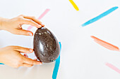 Cropped unrecognizable person's hand gently clasps a large speckled decorative egg against a white surface with vibrant paint strokes.