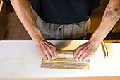 From above of unrecognizable tattooed male chef in apron standing near table and making sushi roll with bamboo mat with hands while working in kitchen