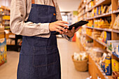 Cropped unrecognizable male worker in casual clothes and apron standing near shelves and working on tablet in modern lighted grocery shop