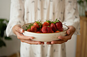 Crop anonymous woman in white gown showing white bowl with fresh strawberries while standing on blurred background in daylight