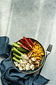 Top view of five ingredients bowl with raw organic vegetables cauliflower, tomato, cucumber, boiled sweet corn and barbeque chicken meat placed on gray surface near napkin and fork