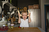 Portrait of cute Caucasian boy with blonde hair showing bananas and smiling at camera while standing and cooking in kitchen at home