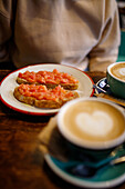 Köstliche, appetitliche Toasts mit Tomaten auf einem Teller neben Tassen mit aromatischem Cappuccino auf einem Holztisch in einer Cafeteria