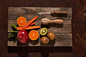Draufsicht auf frische reife Kiwi und Orangen neben Karottenscheiben und Granatapfel, arrangiert mit Messer und Saftpresse auf einem hölzernen Schneidebrett