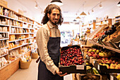 Side view of friendly ethnic male seller with curly hair smiling while showing box of fresh ripe plums standing at counter with assorted fruits and vegetables in supermarket