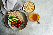 Top view of healthy plant-based plate with hummus and vegetables served in bowls near napkin and glass of liquor against gray background