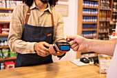 Crop anonymous male worker in casual cloth with apron receiving payment from client with POS machine and credit card while working in drugstore