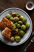 A rustic meal consisting of golden brown, pan-fried brussels sprouts and crispy fish fillets, served on a ceramic plate with a side of chili flakes