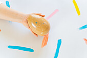 Cropped unrecognizable person's hand gently clasps a large speckled decorative egg against a white surface with vibrant paint strokes.