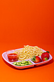 From above of plate of spaghetti with tomato sauce cucumber slices and strawberries served for school lunch on red table
