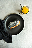 Draufsicht auf ein Sandwich aus Kohlensauerteigbrot, Lachs und Avocadoscheiben zum Frühstück, serviert auf einem schwarzen Teller auf dem Tisch neben einer Serviette und einem Glas Saft mit Löffeln vor grauem Hintergrund