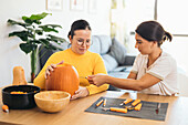 Focused young Hispanic female friends looking down while sitting at table with Halloween kit bowls and carving eye with sharp tool on orange pumpkin