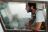 Seitenansicht eines bärtigen hispanischen Mannes im T-Shirt, der wegschaut, während er in der Nähe des Kabinenfensters mit dem Armaturenbrett sitzt und auf dem Meerwasser von Soller Mallorca segelt