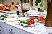 Close-up of a luxurious dining table arrangement showcasing a red flower on a plate, a folded napkin, porcelain dishes with gold detailing, a brass pitcher, and a glass of red wine on a lace tablecloth.