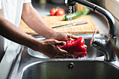 Cropped unrecognizable African American male washing fresh bell pepper in sink with water stream while cooking healthy salad in kitchen