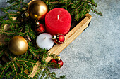 From above of Christmas baubles candles and wooden plank placed on grey surface near green fir branches