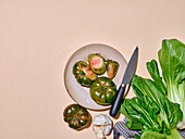 Grüne Tomaten mit anderem Gemüse in hellem Sonnenlicht, flach auf einem Teller liegend, mit einem Messer mit Kopierraum auf beigem Hintergrund