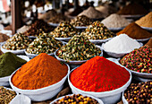 Rows of mix colored powdered spices and seeds in trays at local market stall over blurred busy market background