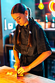 Focused young female chef with braids in black uniform with focus cutting ingredient with knife on chopping board while preparing meal at illuminated counter in bistro