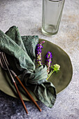 Hoher Winkel eines herbstlich gedeckten Tisches mit marmoriertem Teller, Serviette, Gabel und Messer und Lavendelblüten neben einem leeren Glas vor einer grauen Fläche