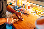 High angle of Crop hands of unrecognizable chef wearing gloves and cutting fresh sushi roll with knife on wooden chopping board at bar counter