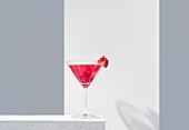 Glass filled with red pomegranate cocktail served with pomegranate seeds against a gray and white backdrop, casting shadow