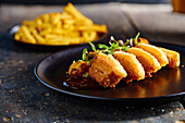 Delicious fried breaded meat garnished with mushrooms and herbs served on black plate near French Fries against blurred background