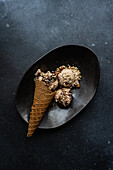 Top view of black ceramic plate with a cone of chocolate ice cream accompanied by two scoops of ice cream with peanuts and walnuts