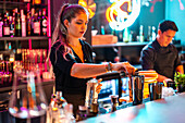 Young beautiful Caucasian barmaid in black uniform pouring liquor from jigger in shaker at illuminated counter in modern nightclub