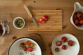 Draufsicht auf frisch geschnittene Erdbeeren auf einem Holzbrett und in einer Schüssel mit Messer auf einem Holztisch bei der Zubereitung von Erdbeermarmelade in einem Haus