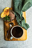 Blick von oben auf ein Glas hausgemachtes Kaffeeeis mit Waffel und frischer Minze neben einem Löffel und einer Tasse Kaffee auf einem Holztablett mit Serviette auf einer grauen Fläche