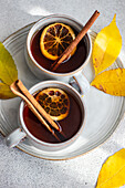 From above of two cups of aromatic spiced tea with cinnamon sticks, anise, and dried orange slices surrounded by radiant yellow autumn leaves on a textured gray background