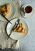 Top view of plates with pieces of apple pie and forks on napkin near cup of tea against gray table