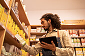 From below side view of focused young ethnic male worker in casual clothes and apron standing near shelves while checking goods in modern grocery shop