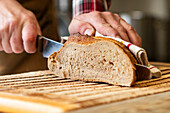 Closeup of crop anonymous person's hands skillfully slicing fresh bread using knife and napkin on a wooden cutting board against blurred background