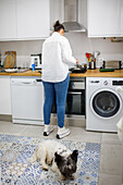 Back view full body of anonymous female in casual outfit cooking food at kitchen counter with cute fluffy dog at home