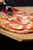 Anonymous crop person sitting at wooden table with delicious pizza and holding stainless steel cutter with hand and cutting slices against dark background