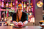 Happy beautiful young barmaid in black uniform smiling at camera while serving fresh cocktail at counter in modern nightclub