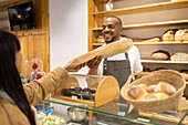 Lächelnder afroamerikanischer Mann in einem Bäckereiladen, der vor Produkten in Regalen und Glasvitrinen steht und einer anonymen Kundin eine Tüte reicht