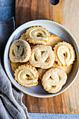 From above fresh homemade baked butter cookies placed in a bowl on a wooden cutting board beside a gray napkin, set against a minimalist concrete background