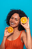 Optimistic young Hispanic female with curly black hair smiling and looking at camera while holding orange slices against blue background