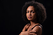 Side view of Hispanic young female with curly hair and crossed arms looking at camera while standing on black background