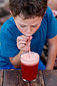 High angle view of little boy in casual clothes drinking fresh watermelon juice in glass with straw while sitting at wooden table in restaurant