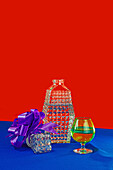 Ein Geschenk mit einer lila Schleife neben einer Kristallvase auf einer blauen Fläche mit einem kräftigen roten Hintergrund