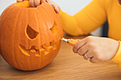 Anonymous female sitting at table and carving with tool scary teeth on orange pumpkin and preparing for Halloween decoration in day