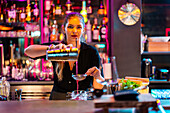 Beautiful confident young female barkeeper looking at camera and straining drink from shaker at bar counter while working in nightclub